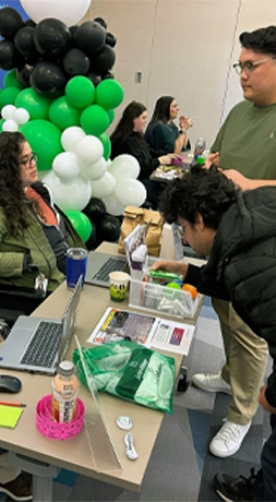 テーブル、ノート PC、情報資料が並ぶ屋内学習フェアや博覧会に参加する人々。