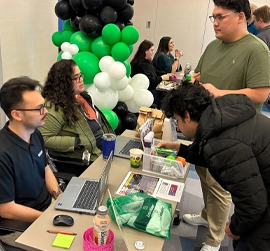 テーブル、ノート PC、情報資料が並ぶ屋内学習フェアや博覧会に参加する人々。