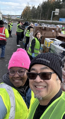 黄色の安全ベストを着た二人が、屋外のボランティアイベントで笑顔で自撮りをしている。