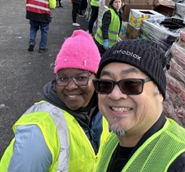 黄色の安全ベストを着た二人が、屋外のボランティアイベントで笑顔で自撮りをしている。