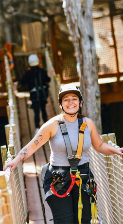 安全装備を身に着けた笑顔の女性が、屋外でのアドベンチャー活動中に、ロープの橋の上に立っている。