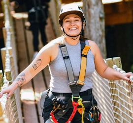 安全装備を身に着けた笑顔の女性が、屋外でのアドベンチャー活動中に、ロープの橋の上に立っている。