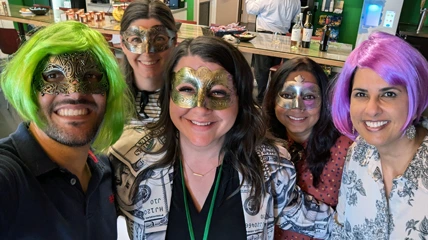 お祝い用のマスクとカラフルなかつらをつけた5人が、オフィスの祝賀会で楽しいグループセルフィーを撮っている。