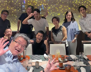 レストランの長いテーブル越しに人々が笑顔で手を振っている楽しい夕食会。