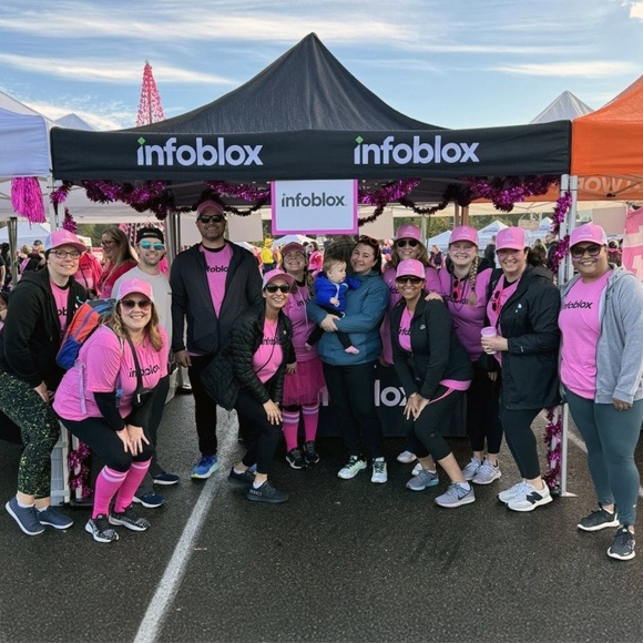 ピンクの服を着た Infoblox のチームメンバーが、屋外のチャリティーイベントで装飾されたブースの前でポーズをとっている。